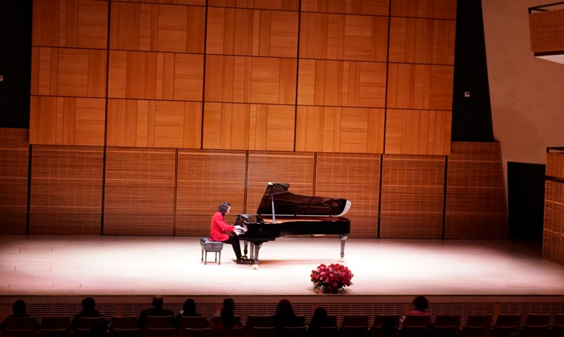 A pianist performing on a grand piano on stage in a concert hall, with a small audience watching.